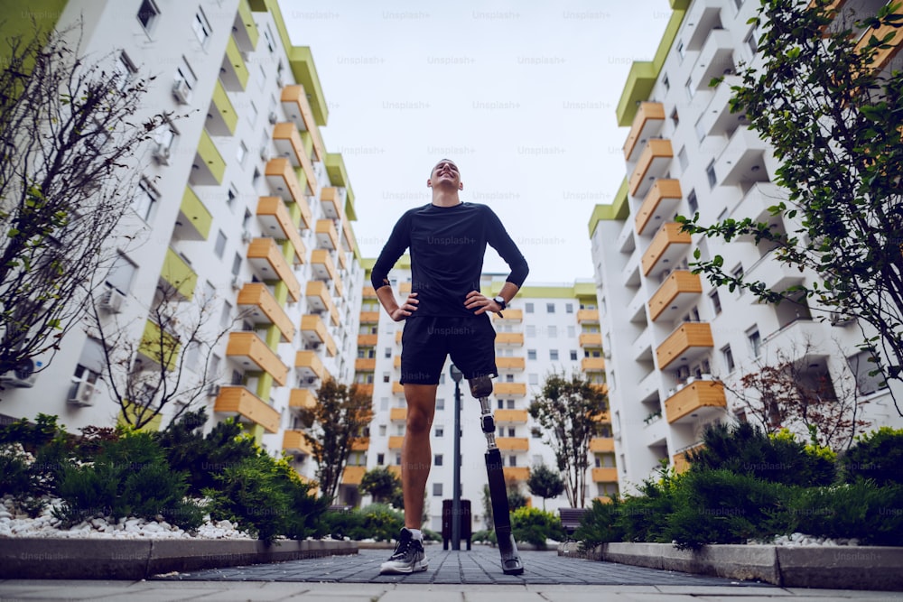 Vue en contre-plongée d’un beau sportif avec une jambe artificielle debout avec les mains sur les hanches à l’extérieur entouré de bâtiments.