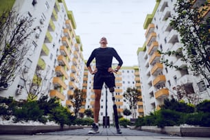 건물로 둘러싸인 야외에서 엉덩이에 손을 얹고 서 있는 인공 다리를 가진 잘생긴 스포츠맨의 낮은 각도 보기.