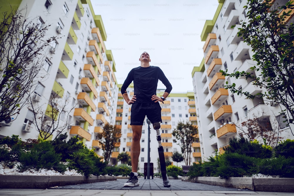 Visão de baixo ângulo de um esportista bonito com perna artificial em pé com as mãos nos quadris ao ar livre cercado por edifícios.