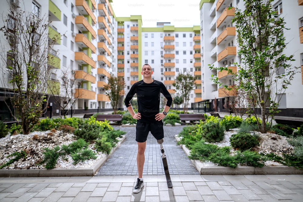 In voller Länge eines gutaussehenden lächelnden Sportlers mit künstlichem Bein, der mit den Händen in den Hüften im Freien steht, umgeben von Gebäuden. Rücklicht.