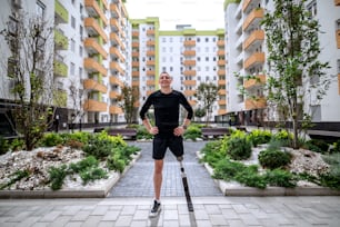 Comprimento completo de esportista sorridente bonito com perna artificial em pé com as mãos nos quadris ao ar livre cercado por edifícios. Luz de fundo.