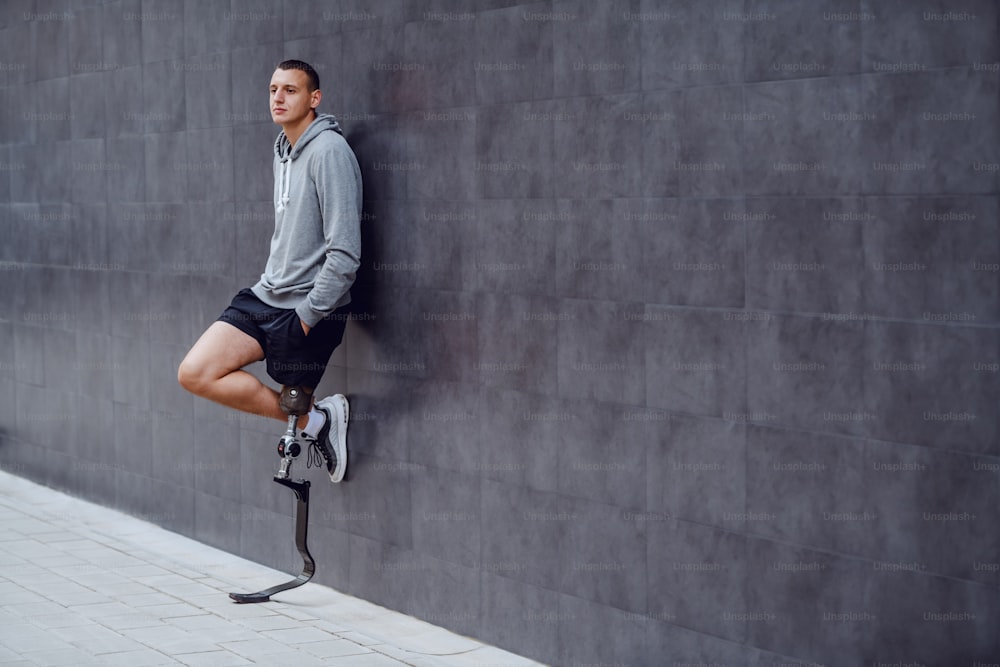 Full length of handsome caucasian sportsman with artificial leg leaning on the wall outside and having hands in pockets.