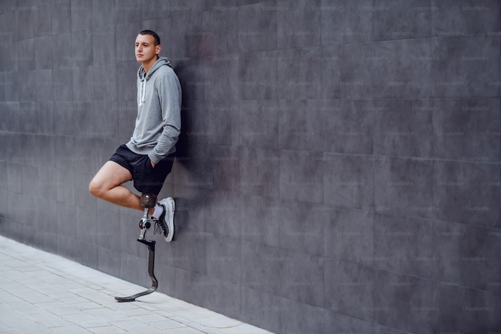 Cuerpo entero de apuesto deportista caucásico con pierna artificial apoyada en la pared exterior y con las manos en los bolsillos.