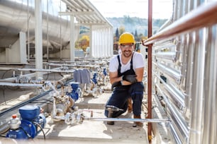 Handsome caucasian unshaven worker in working clothes and with helmet on head kneeling next to oil tank and looking at camera.