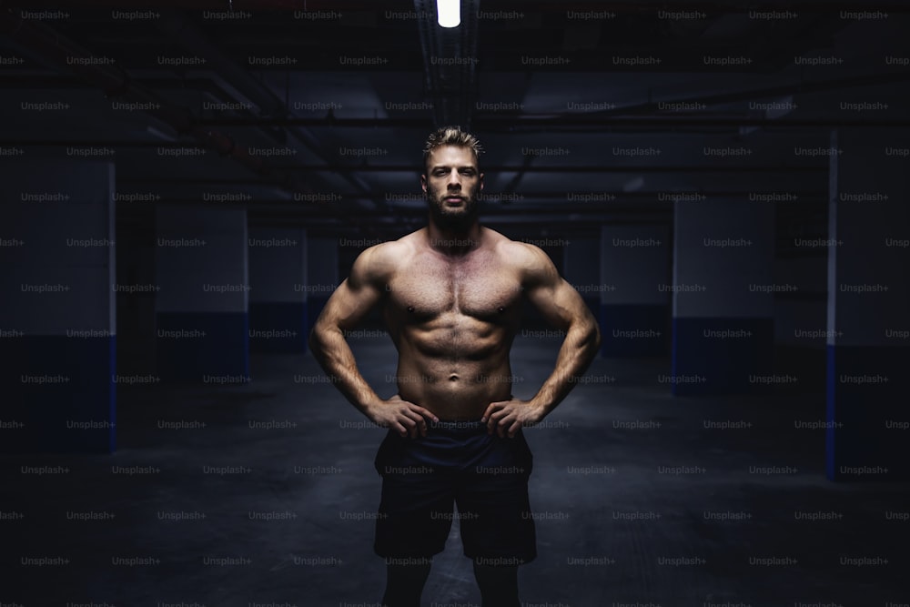 Three quarter length of topless muscular sportsman shorts standing in garage with hands on hips at night and looking at camera. Urban life concept.