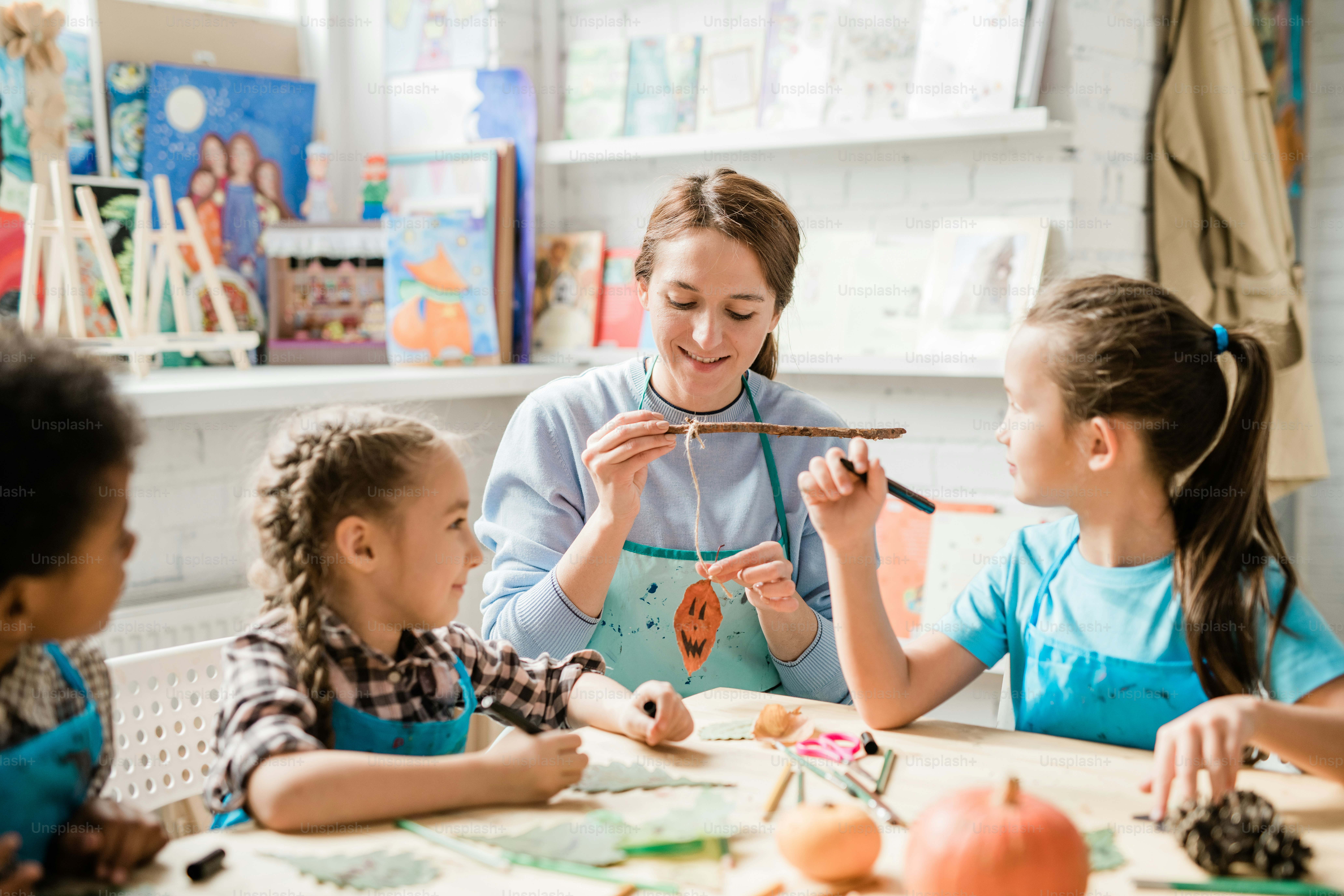 Happy young teacher holding halloween pumpkin decoration on thread while showing group of schoolkids how to make it