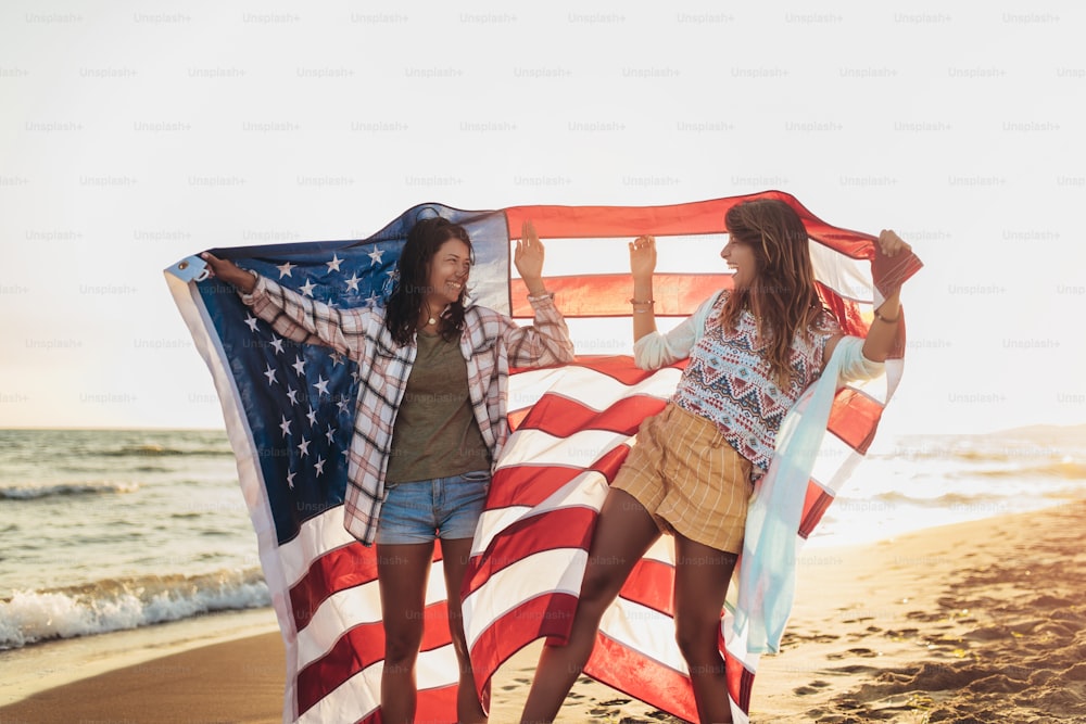 Felices amigas jóvenes disfrutan de un día soleado en la playa. Están sosteniendo una bandera estadounidense.