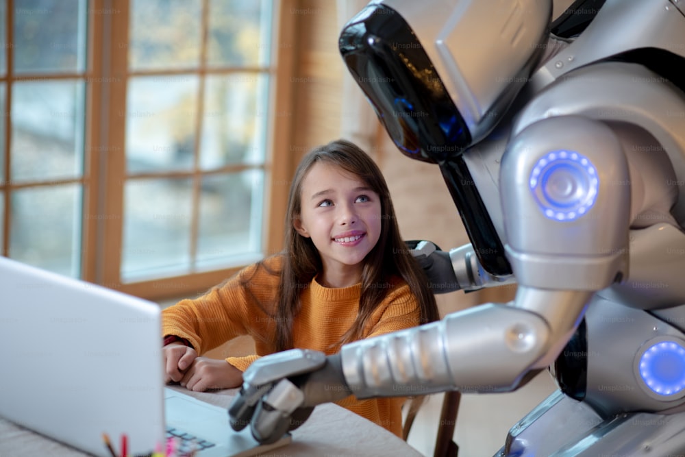 Lets do it together. Robot helping a girl with her homework showing her something on laptop