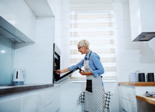Side view of attractive caucasian senior blond woman in apron standing in kitchen and taking out baked dish from oven.