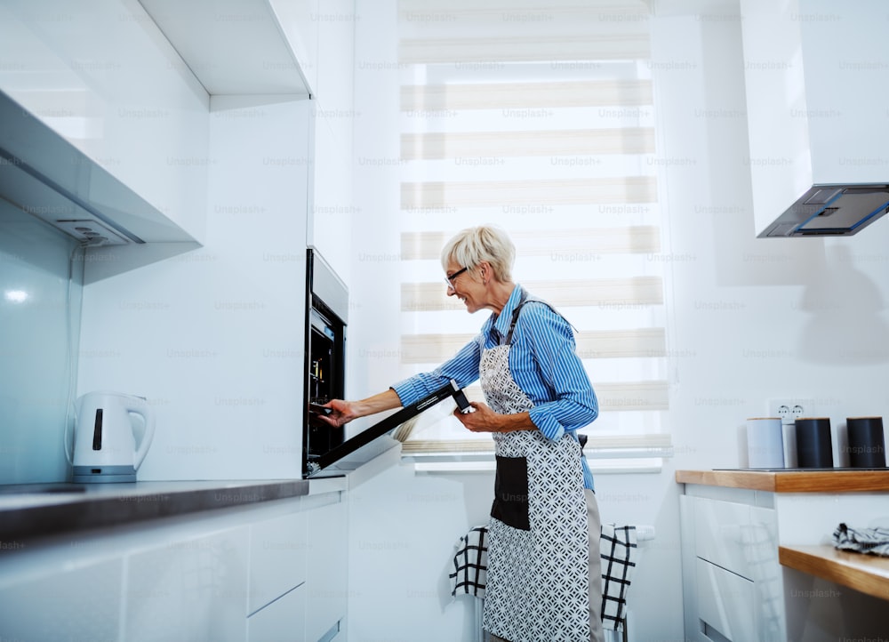 Side view of attractive caucasian senior blond woman in apron standing in kitchen and taking out baked dish from oven.