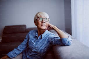 Portrait of smiling charming caucasian blond woman sitting on sofa in living room next to window and looking trough it.
