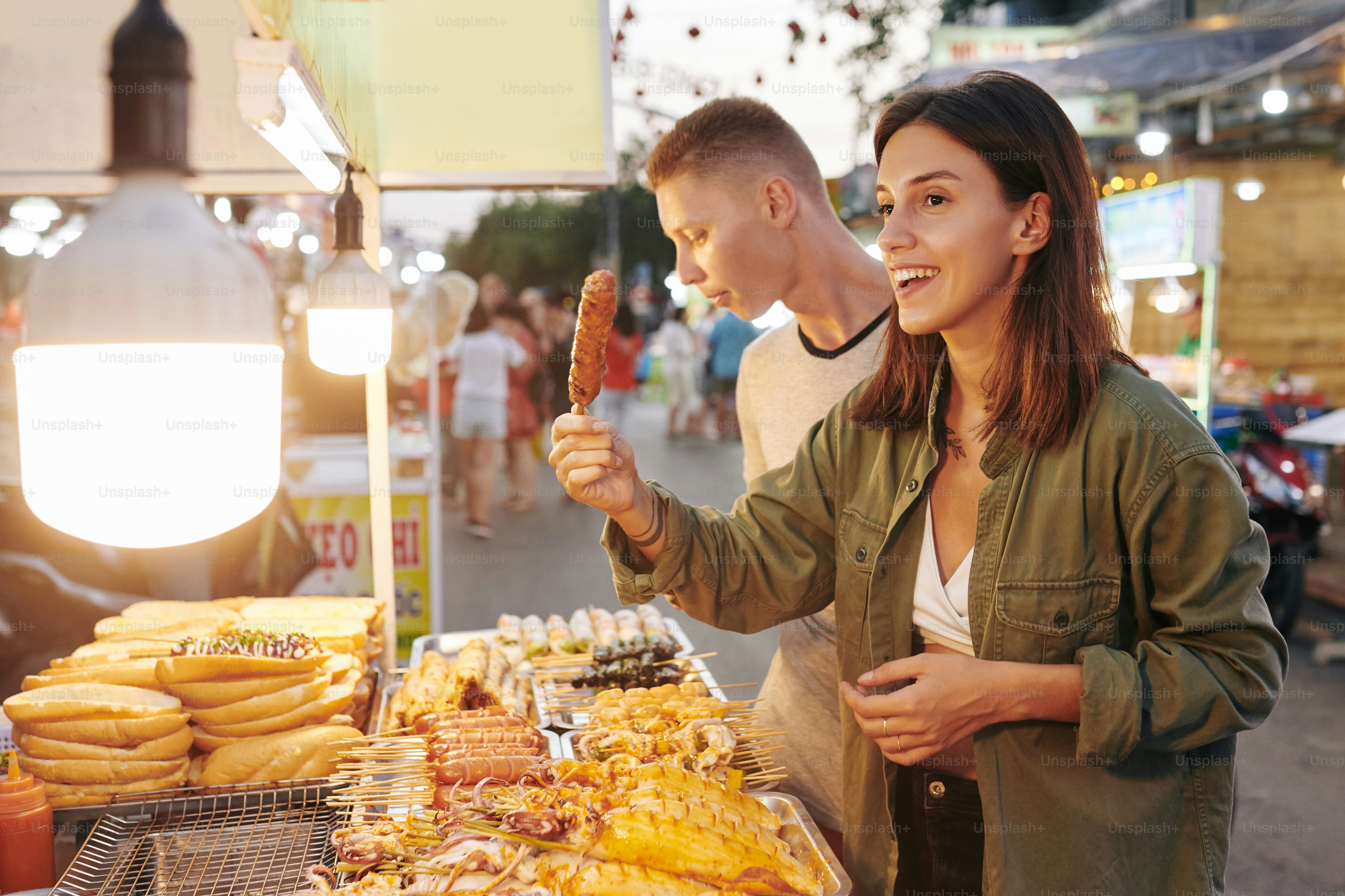 Happy young woman buying grilled pork skewer at stall with traditional Vietnamese street food