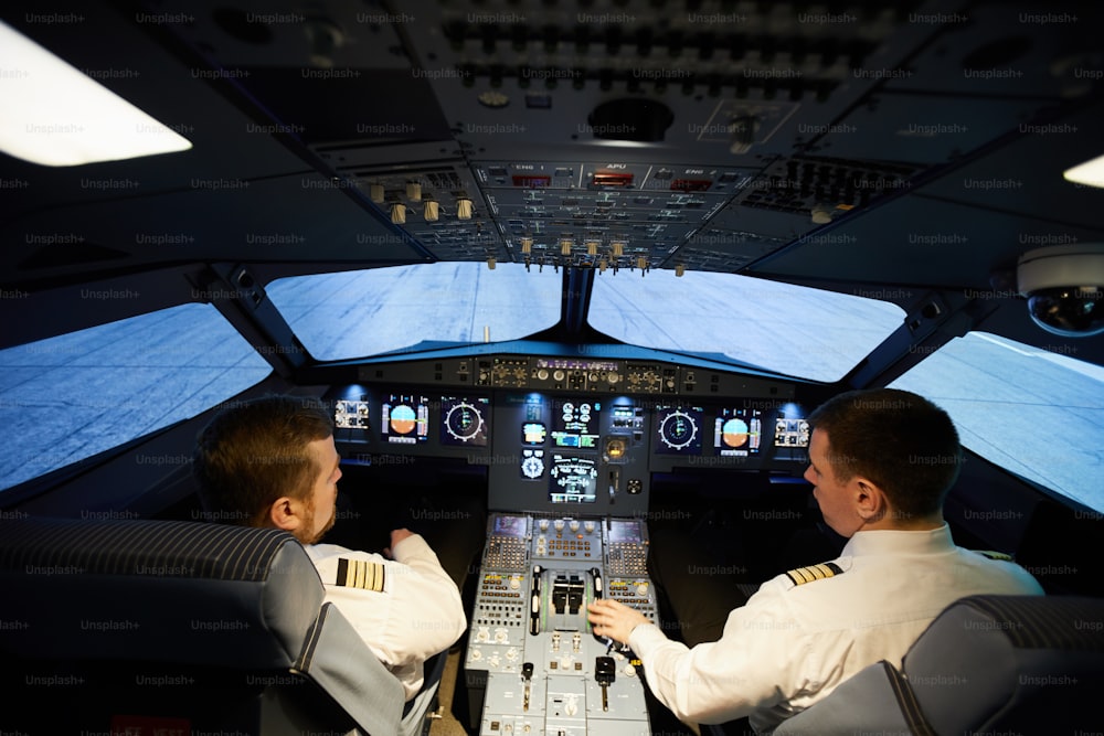 Rear view of serious concentrated pilots in white shirts with stripes sitting at control panel of aircraft and preparing for flight