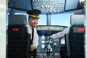 Portrait of smiling handsome bearded pilot in captains cap sitting in leather armchair and turning back in cockpit