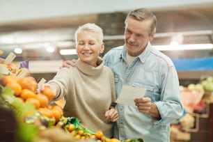 Waist up portrait of modern senior couple choosing fruits while enjoying grocery shopping at farmers market, copy space