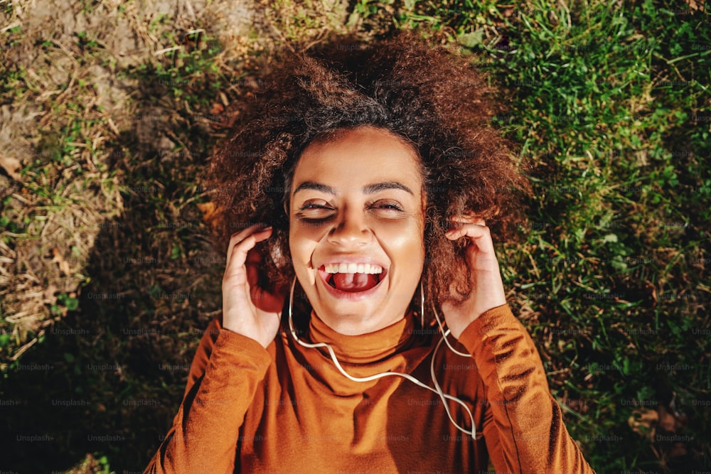Top view of beautiful smiling cheerful young mixed race woman with curly hair lying down on lawn and listening music over headphones.