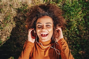 Vista superior de la hermosa joven sonriente y alegre mujer de raza mixta con cabello rizado acostada en el césped y escuchando música con auriculares.