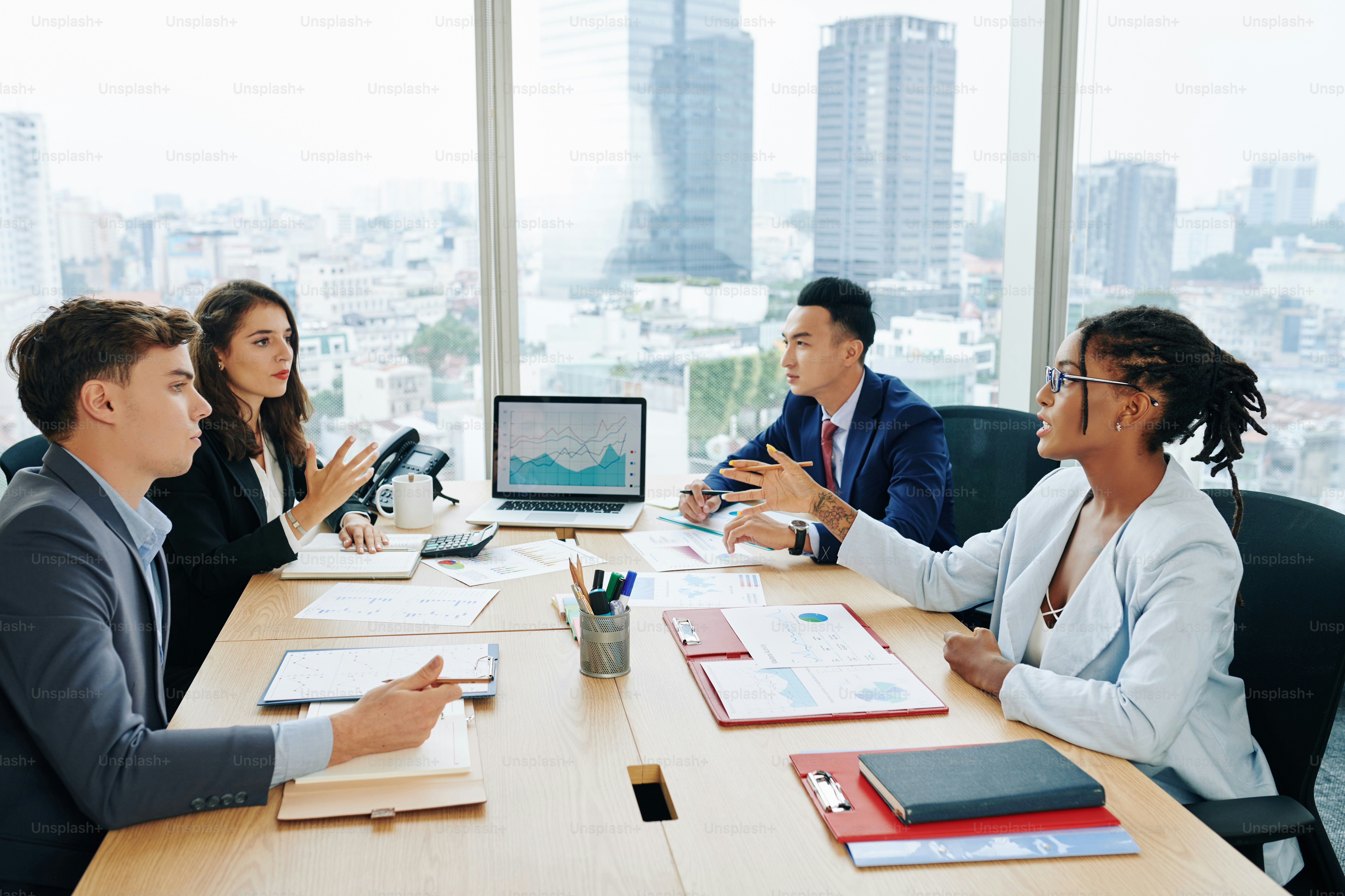 Multi-ethnic business team sitting at big office table and discussing sales report and plans at meeting