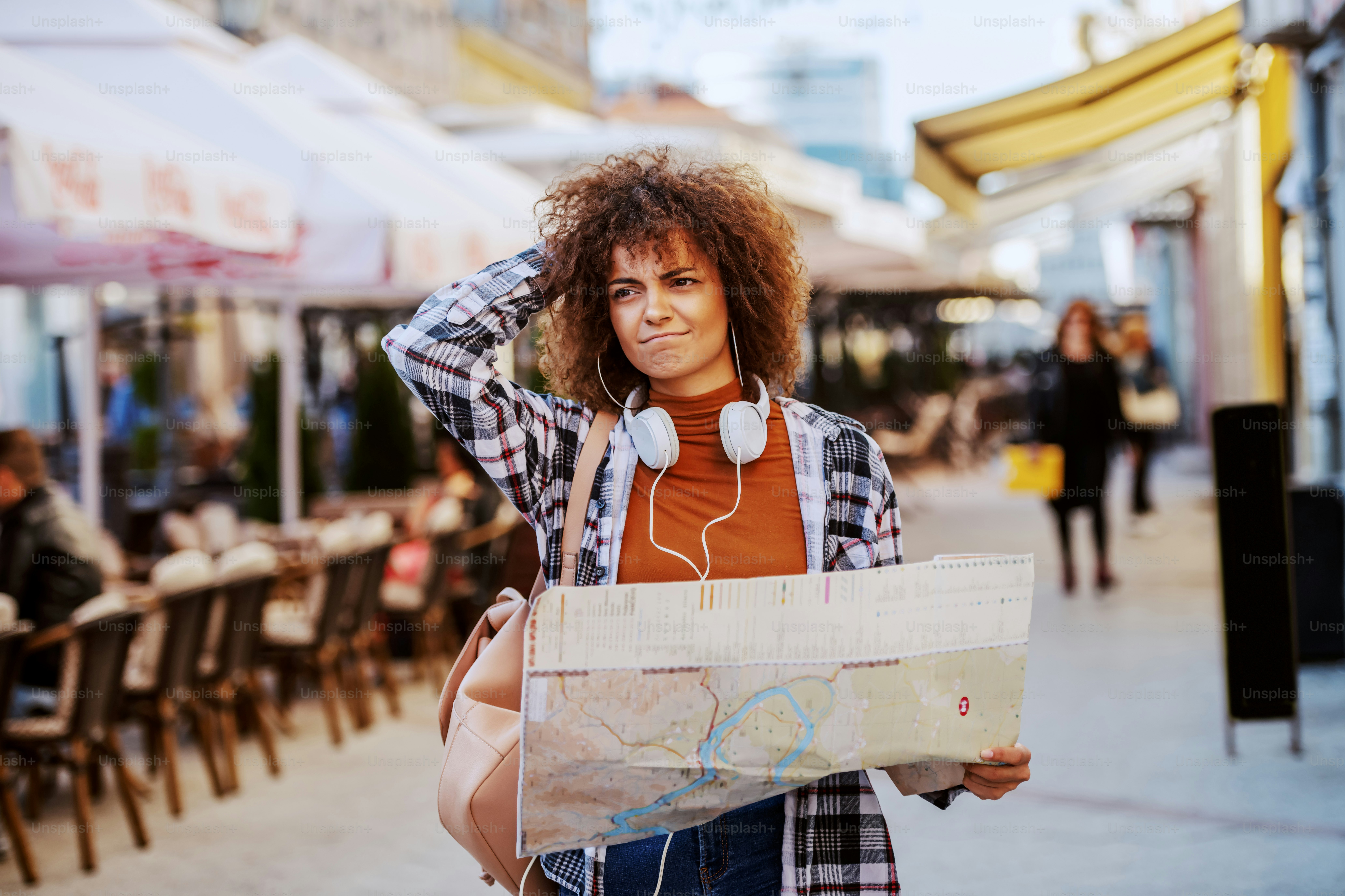 Verlorene junge gemischtrassige Touristin mit lockigem Haar und in kariertem Hemd, die einen Stadtplan hält und sucht, wohin sie gehen soll, während sie in der Hauptstraße steht.