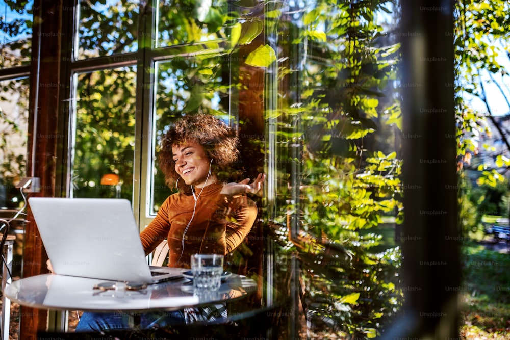 Hermosa joven sonriente mujer de raza mixta con cuello alto y con cabello rizado sentada en una cafetería y teniendo videollamada a través de la computadora portátil. En los oídos hay auriculares. Foto tomada desde el exterior.