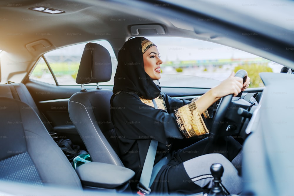 전통 의상을 입은 젊은 화려한 웃는 무슬림 여성이 값비싼 차를 운전하고 있다.