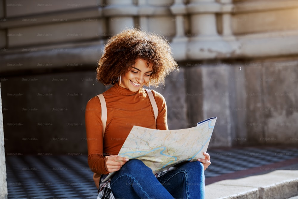 Bela jovem turista mestiça feminina com cabelo encaracolado sentado na rua da cidade e olhando para o mapa.