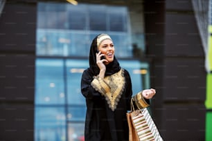Splendida donna musulmana sorridente positiva attraente in abiti tradizionali in piedi di fronte al centro commerciale con borse della spesa in mano e taxi di chiamata.