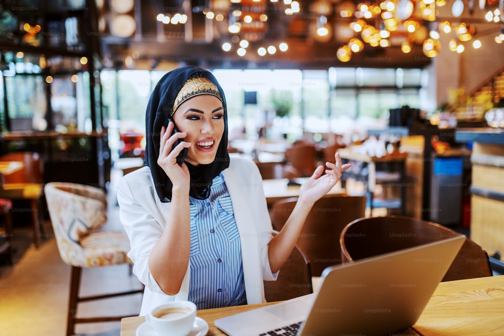 Charmante jeune femme d’affaires musulmane à la mode assise dans un café et parlant au téléphone avec le patron. Sur la table se trouvent du café et un ordinateur portable. Génération du millénaire.