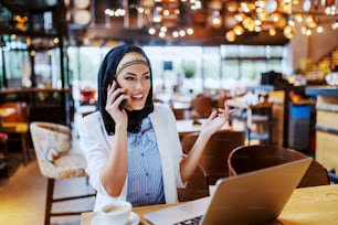 Lovely young muslim fashionable businesswoman sitting in cafe and talking on the phone with boss. On table are coffee and laptop. Millennial generation.