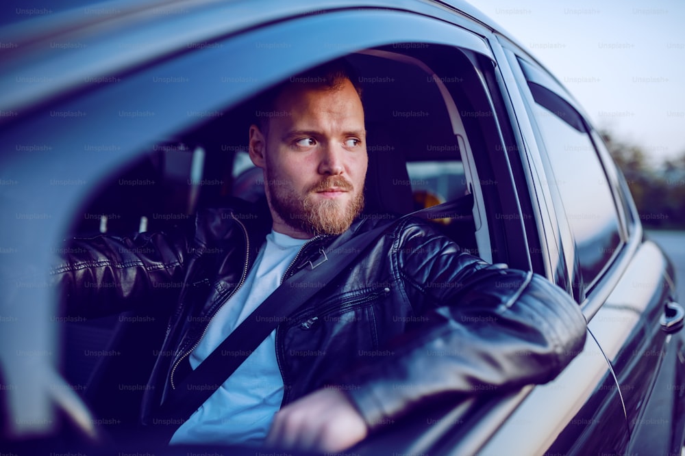Young handsome pensive bearded blond caucasian man in leather jacket posing in his modern car.