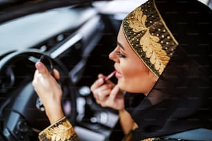 Hermosa y atractiva joven musulmana con ropa tradicional sentada en su automóvil durante el atasco de tráfico y poniéndose lápiz labial.