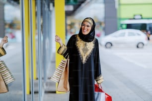 Atraente mulher árabe sorridente em traje tradicional olhando para a vitrine enquanto está de pé com sacolas de compras nas mãos.