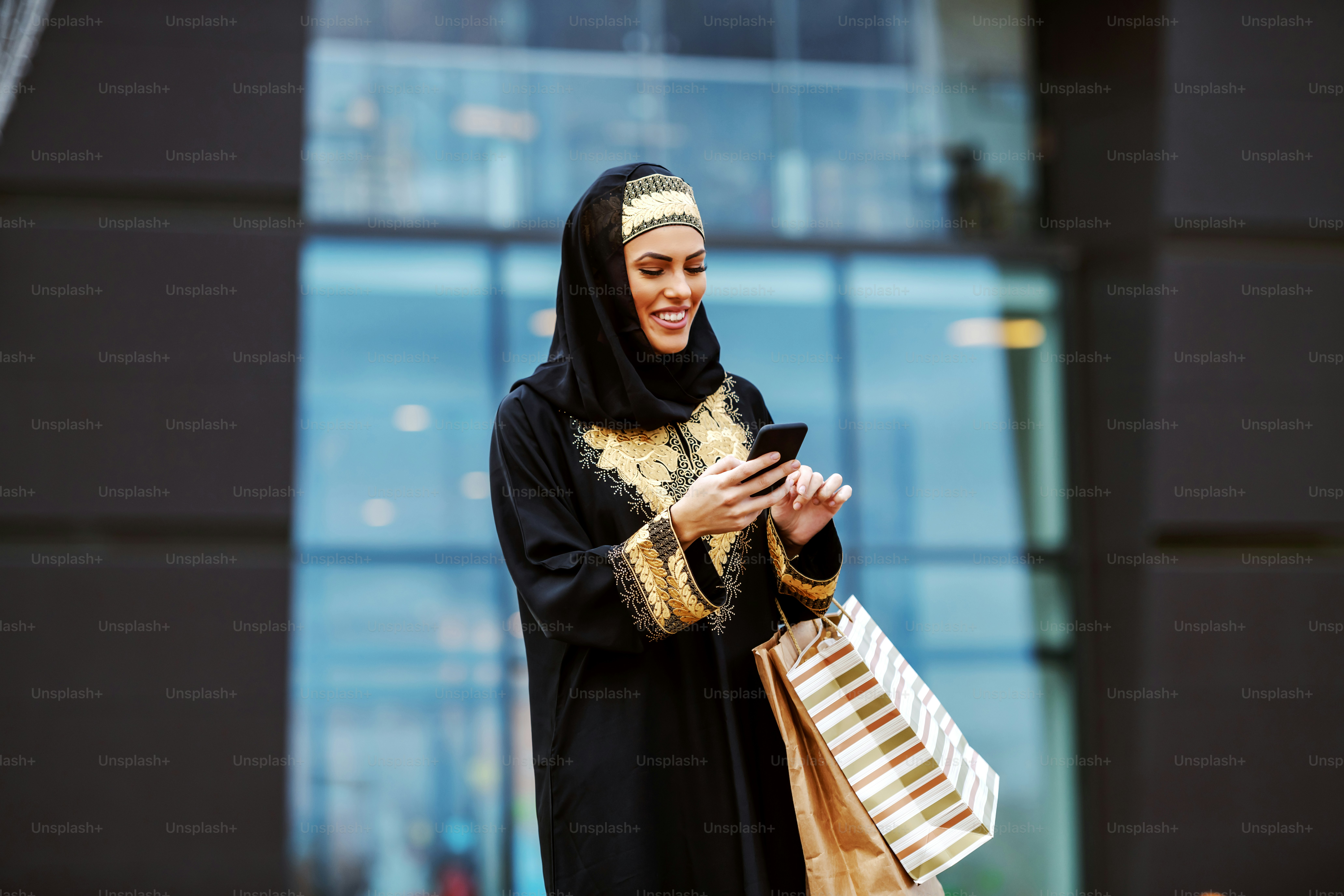 Attractive arab woman in traditional wear standing in front of shopping mall with shopping bags in hands and using smart phone for reading or sending message.