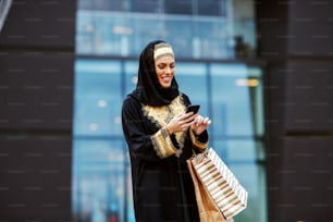 Mulher árabe atraente em traje tradicional em pé em frente ao shopping center com sacolas de compras nas mãos e usando o telefone inteligente para ler ou enviar mensagem.