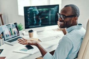 Hübscher junger afrikanischer Mann im Hemd, der den Computer benutzt und lächelt, während er im Büro arbeitet