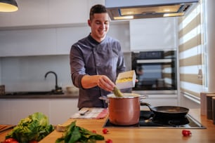 Jovem feliz sorridente chef caucasiano em uniforme colocando macarrão na panela com água fervente. Cozinha italiana.
