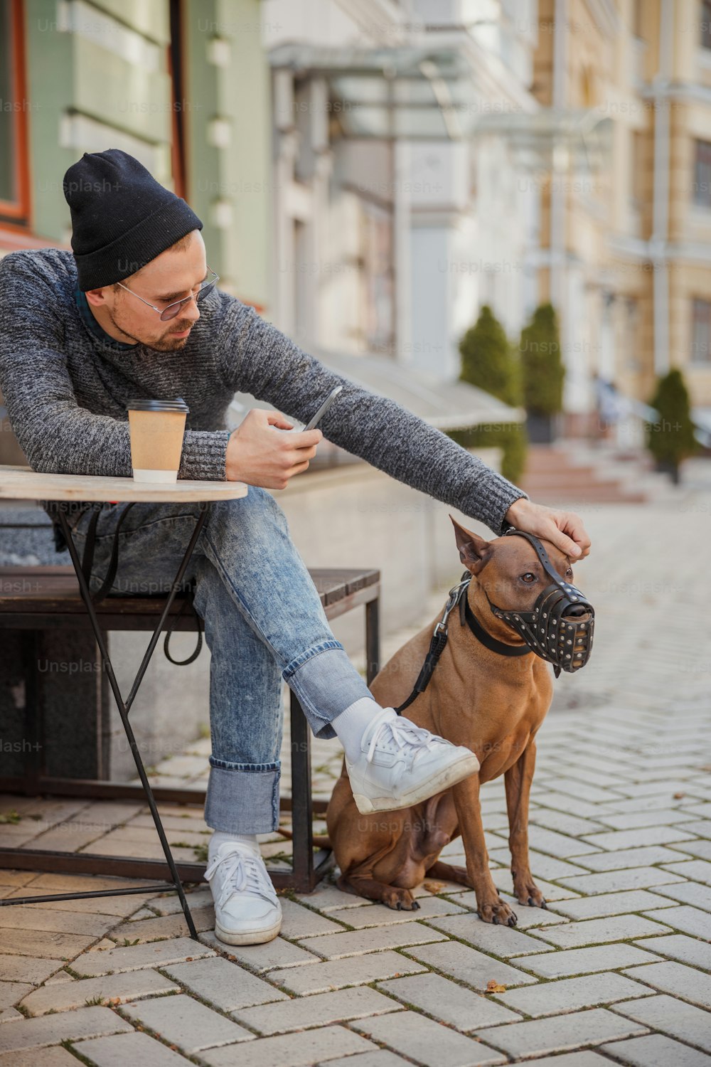 Junger Besitzer mit Smartphone sitzt mit seinem großen Hund am Tisch im Freien