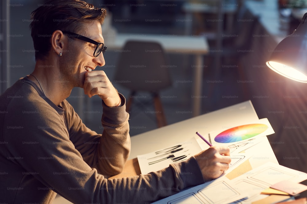 Side view portrait of contemporary male designer drawing sketches at desk while working in studio lit by lamp light, copy space