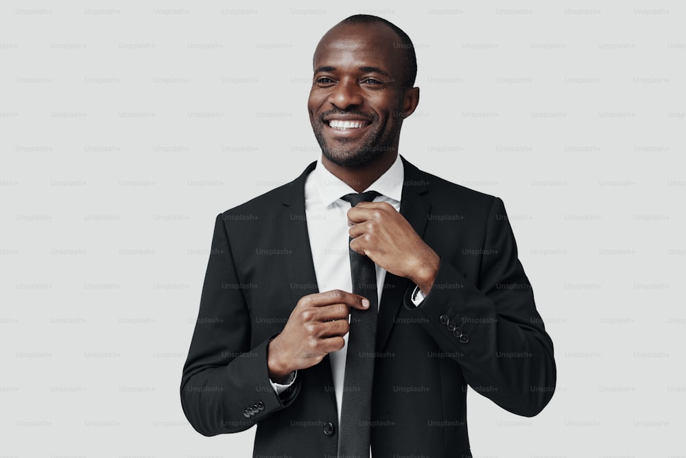 Elegante joven africano con ropa formal ajustándose la corbata y sonriendo mientras está de pie sobre un fondo gris