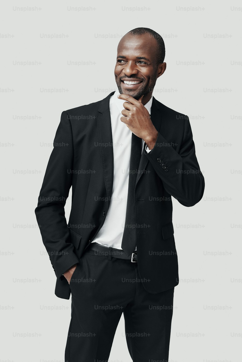Élégant jeune homme africain en tenue de cérémonie regardant ailleurs et souriant tout en se tenant sur un fond gris