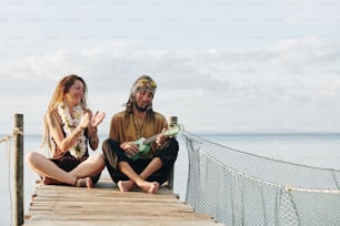 Cheerful pretty mature woman with flower necklace clapping to her husband playing ukulele when thay are sitting on pier