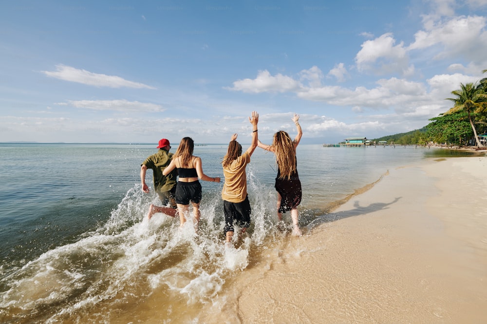 Vista trasera de jóvenes felices y emocionados corriendo y chapoteando en el agua en la playa