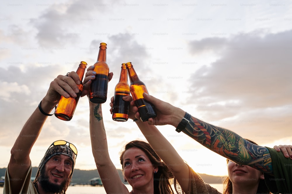Amigos alegres brindando y tintineando botellas de cerveza cuando tienen fiesta en la playa