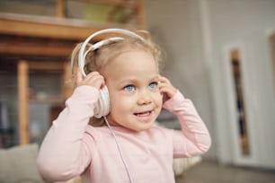 Cintura para cima retrato de menina bonito de olhos azuis usando fones de ouvido grandes e apreciando a música no interior de casa, espaço de cópia