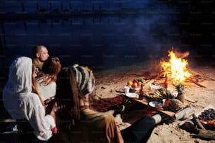 Friends resting on beach in the evening, they are sitting by fire and eating fresh fruits and seafood