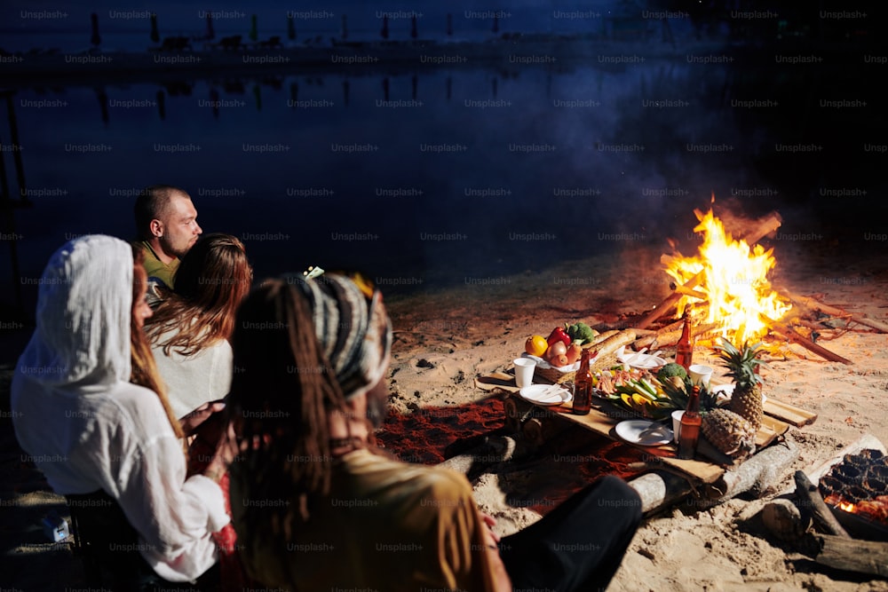 Friends resting on beach in the evening, they are sitting by fire and eating fresh fruits and seafood