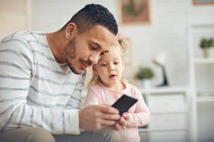 Retrato de un padre contemporáneo que muestra un teléfono inteligente a una linda hija pequeña en casa, copie el espacio