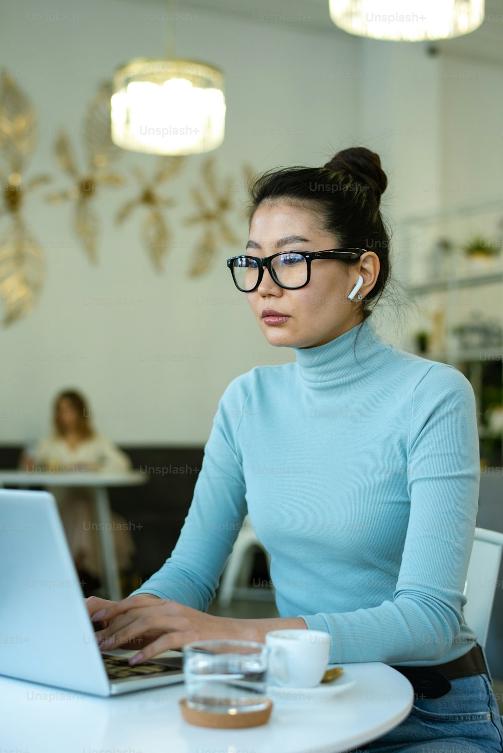 Mujer joven y seria con auriculares inalámbricos sentada frente a la computadora portátil junto a la mesa y mirando la información en línea