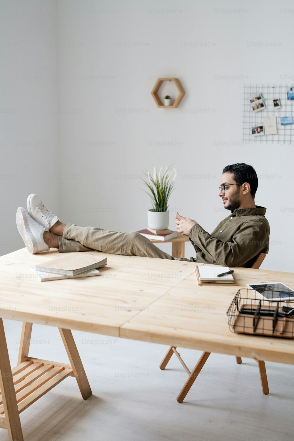 Young office employee in casualwear scrolling in smartphone while sitting by workplace with his legs on table