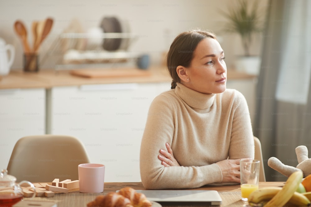 Warm toned portrait of elegant adult woman looking at window while enjoying breakfast in cozy kitchen, copy space